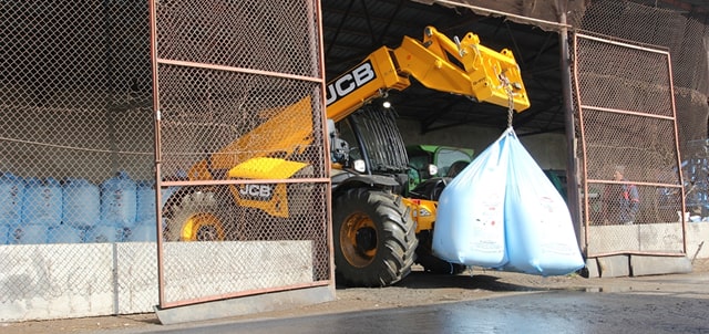 A yellow JCB telehandler lifting two large blue bags out of a fenced enclosure.