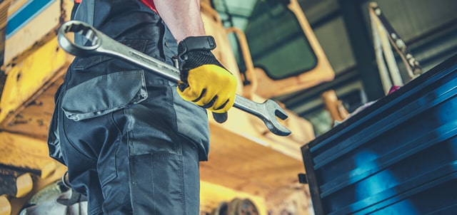 Man in overalls holding a wrench near a vehicle.